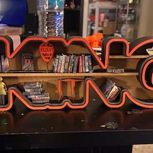 May include: A black and orange wooden shelf shaped like the word "KING" with a red balloon that says "All work and no play makes Jack a dull boy" and a paper boat on the top shelf. The shelves are filled with miniature books and figures.
