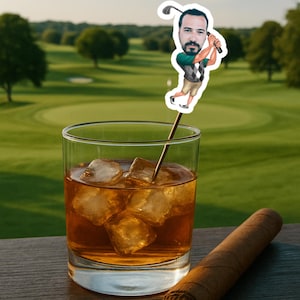 May include: A rocks glass filled with ice and a dark amber liquid, with a golf-themed cocktail pick featuring a cartoon golfer. A cigar rests on a wooden surface, with a golf course in the background.