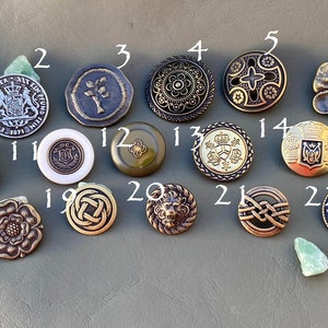 May include: Twenty-six assorted metal buttons with various designs, including floral, geometric, and heraldic motifs. The buttons are arranged in rows on a gray surface.
