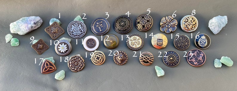 May include: Twenty-six assorted metal buttons with various designs, including floral, geometric, and heraldic motifs. The buttons are arranged in rows on a gray surface.