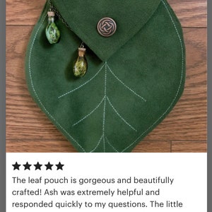 May include: A green leaf-shaped pouch with a flap closure and a decorative metal clasp. Two small glass bottles with cork stoppers and green accents hang from the pouch.