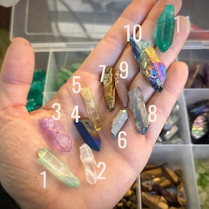 May include: A hand holding ten different crystal points, numbered 1 through 10. The crystals are various colors, including clear, green, purple, gold, and black.
