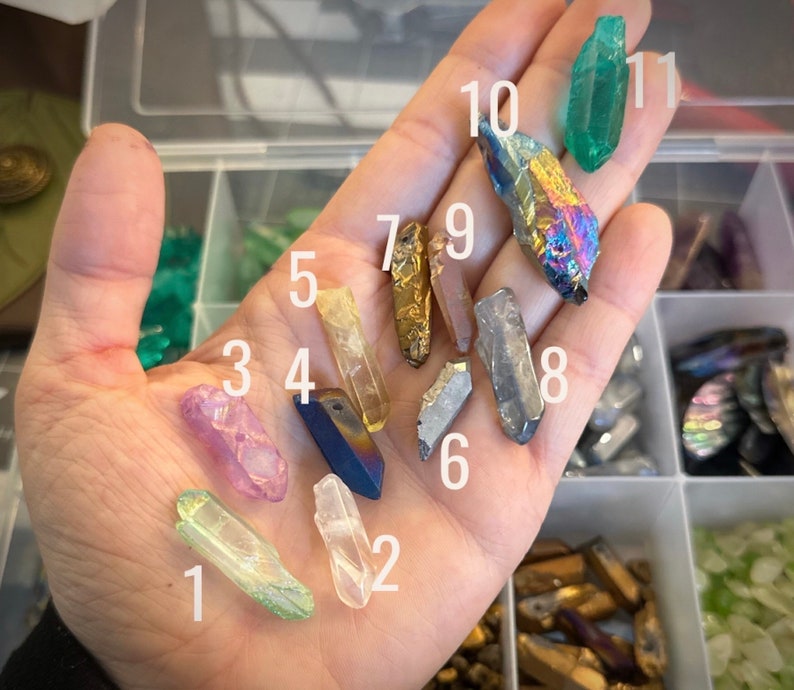May include: A hand holding ten different crystal points, numbered 1 through 10. The crystals are various colors, including clear, green, purple, gold, and black.
