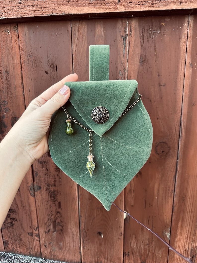 May include: A green velvet pouch shaped like a leaf with a metal clasp and a chain with two small glass vials hanging from it.