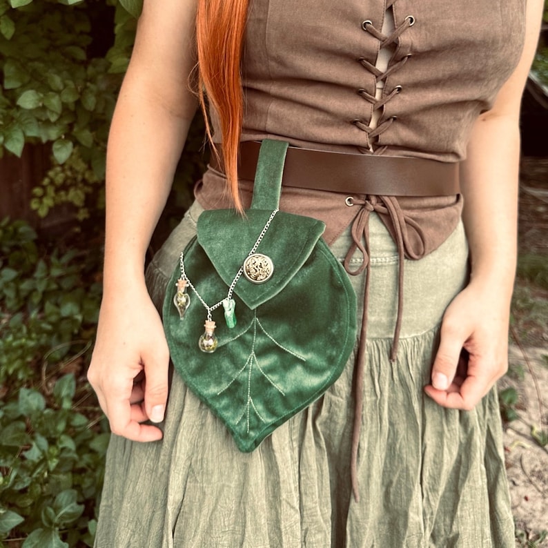 May include: A green velvet leaf-shaped pouch with a metal button and chain. The pouch is attached to a brown leather belt with a metal buckle. The pouch is decorated with small glass vials.