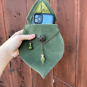 May include: A green velvet leaf-shaped pouch with a phone inside. The pouch has a chain with two small glass vials filled with green plant material.