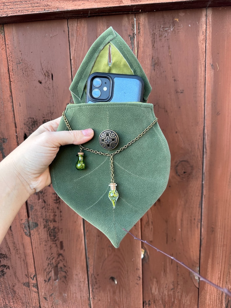 May include: A green velvet leaf-shaped pouch with a phone inside. The pouch has a chain with two small glass vials filled with green plant material.