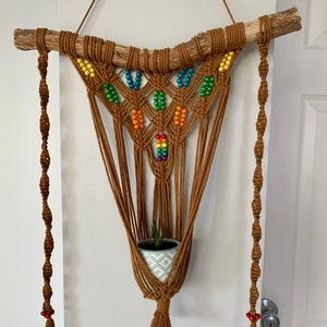 Puede incluir: Un macetero de macramé con una rama de madera marrón en la parte superior y una pequeña planta en maceta colgando en el centro. El macramé está hecho con cordón marrón y tiene cuentas de colores arcoíris tejidas en el diseño.