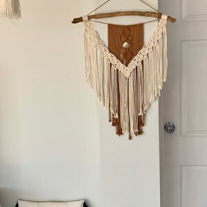 May include: A white pillow with black text that reads "welcome to our nest" sits on a woven brown basket. A small green Buddha statue stands on the floor next to the basket. A macrame wall hanging with a brown and white design hangs on a white wall.