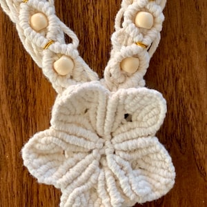 May include: Close-up of a cream-colored macrame necklace with a large flower pendant. The necklace features a series of flower-shaped knots with beige beads and gold accents. The background is a wooden surface.