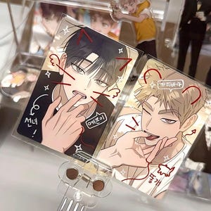 May include: Two clear acrylic fans featuring anime-style illustrations of male characters. One has black hair and the other has blonde hair. Each fan has Korean text and red accents. The fans are held by a clear plastic handle.