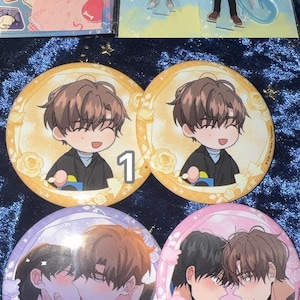 May include: A collection of anime-style merchandise, including character standees, stickers, and round pinback buttons. The buttons feature illustrations of two male characters in various poses, with numbers 1, 2, and 3 on them. The color palette includes pink, blue, and yellow.