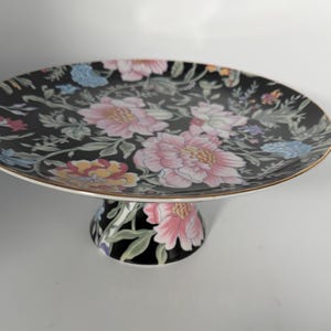 May include: A black ceramic pedestal cake stand featuring a vibrant floral pattern with pink, yellow, and blue flowers and green leaves. The circular plate has a thin gold-toned rim and is supported by a matching flared base with floral motifs.