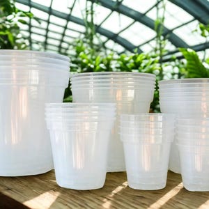 May include: A collection of clear, translucent plastic plant pots arranged in varying sizes. The pots are cylindrical with a slightly tapered base and a wider rim, stacked in groups to show different heights and diameters for gardening needs.