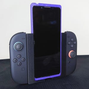 Könnte beinhalten: Ein schwarzes Smartphone wird in einer lila-schwarzen Gamecontroller-Halterung gehalten. Die Halterung hat zwei schwarze Controller-Griffe mit Tasten und Joysticks. Der Telefonbildschirm ist schwarz.