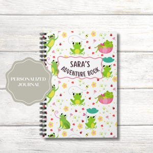 Peut inclure: Un journal à spirales avec une couverture blanche ornée d'un motif répété de grenouilles vertes, de fleurs, de coccinelles et d'abeilles. Le texte "Sara's Adventure Book" est affiché dans une bannière rose et blanche. Un autocollant gris indique "Personalized Journal".