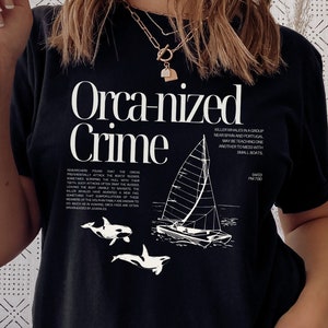 May include: Black t-shirt with the words "Organized Crime" in a bold, white font. The design includes a sailboat illustration and two orca whales. Additional text is present below the main text and illustration.