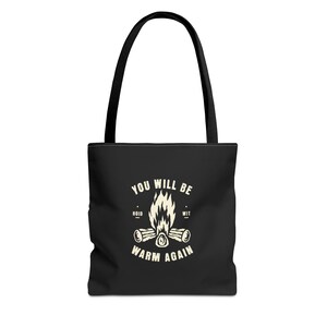 Könnte beinhalten: Schwarze Tragetasche mit schwarzen Griffen. Die Tasche zeigt eine Lagerfeuergrafik mit dem Text "YOU WILL BE WARM AGAIN". Die Grafik ist in Weiß gehalten und zeigt ein Lagerfeuer mit Holzscheiten und Flammen.