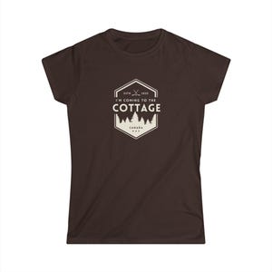 Op de afbeelding: Bruin t-shirt met een witte en beige print. De print luidt "I'M COMING TO THE COTTAGE" met "ESTD 2023" en "CANADA" in een zeshoek met bomen. Een casual t-shirt voor cottage liefhebbers.