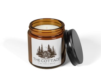 May include: A brown glass jar candle with a black lid. The label reads "THE COTTAGE" above an illustration of a cabin in the woods. The text "STAY FOR A WHILE" is below the illustration. The candle is on a white surface.