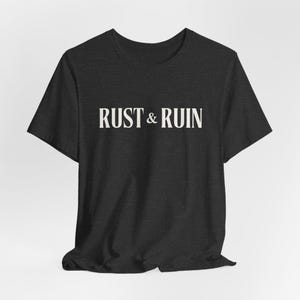 Può includere: T-shirt grigio scuro con la scritta bianca "RUST & RUIN" stampata sul petto. La maglietta ha maniche corte e girocollo. Il tessuto sembra essere un materiale morbido e mélange.
