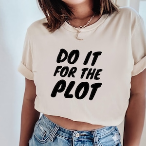 May include: A cream-coloured t-shirt featuring the text "DO IT FOR THE PLOT" in bold, black lettering. The shirt has short sleeves and a crew neck. The model is wearing blue jeans.