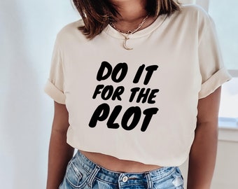 May include: Cream-colored t-shirt with the phrase "DO IT FOR THE PLOT" in bold, black letters. The shirt has short sleeves and a crew neck. The model is wearing blue jeans.