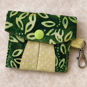 Può includere: Un piccolo portafoglio in tessuto fatto a mano, verde e giallo. Il portafoglio ha una chiusura a patta con un bottone verde e un interno a pois gialli. È attaccato un piccolo cinturino giallo con chiusura in metallo.