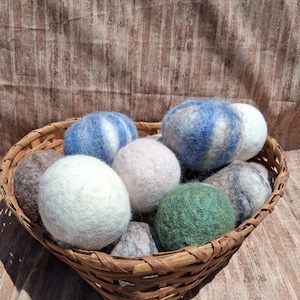 May include: A collection of round, felted wool dryer balls in various colours including white, blue, green, and grey, contained within a woven brown wicker basket. The spheres have a soft, textured surface and are arranged in a cluster inside the basket.