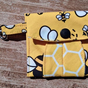 Puede incluir: Una pequeña bolsa de tela amarilla con un patrón repetitivo de abejas en blanco y negro y contornos de panal blancos. La bolsa tiene una solapa plegable asegurada con un botón a presión negro y un mosquetón de metal plateado adjunto.