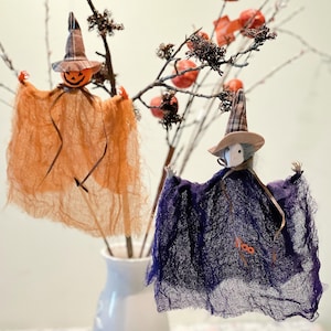 May include: Two hanging ghost decorations with pointed hats, one orange with a jack-o'-lantern face and one purple with a white face, both draped in textured cheesecloth fabric, suspended from thin branches in a white vase.