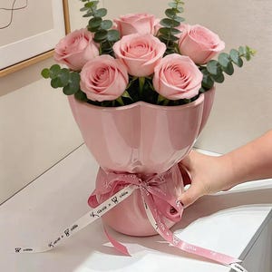 May include: A bouquet of pink roses and green eucalyptus leaves in a light pink ceramic vase. The vase is tied with a pink ribbon with white text. The arrangement is on a white surface.