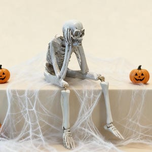 May include: A white skeleton figurine with dark shading, depicted in a seated position with one hand resting against its chin. The skeleton has articulated limbs connected by small metal rings and is positioned between two small orange jack-o'-lanterns.