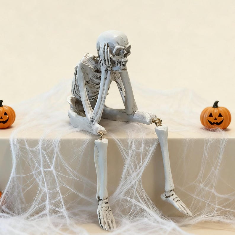 May include: A white skeleton figurine with dark shading, depicted in a seated position with one hand resting against its chin. The skeleton has articulated limbs connected by small metal rings and is positioned between two small orange jack-o'-lanterns.