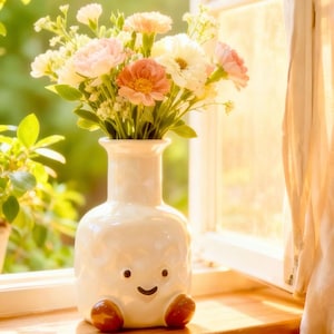 May include: A white ceramic vase with a smiling face and brown feet holds a bouquet of pink, peach, and white flowers. The vase sits on a wooden windowsill, with a window and sheer curtain in the background.
