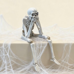 May include: A white skeleton figurine with black detailing, depicted in a seated position with its hands covering its mouth. The skeleton features articulated joints at the hips and knees, allowing the legs to hang down, and has a cracked skull design.