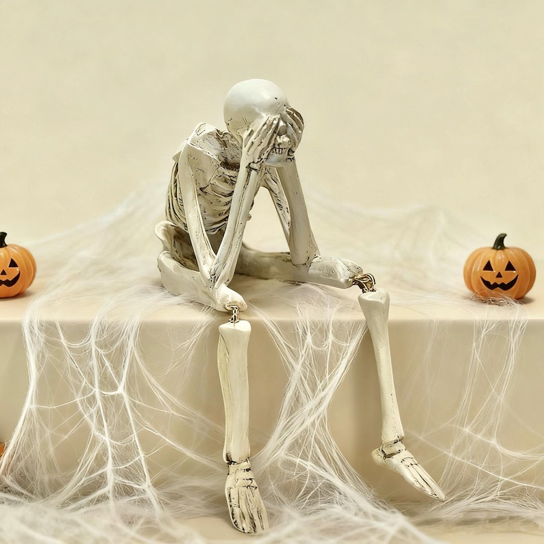 May include: A cream-colored skeleton figurine in a seated position with hands covering its face. The skeleton has a weathered, textured finish and is flanked by two small orange jack-o'-lanterns on a surface draped with white artificial spider web.
