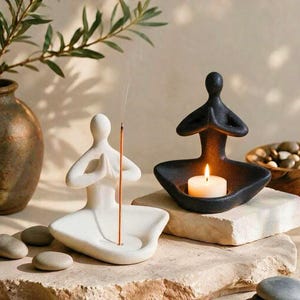 May include: Two ceramic yoga figures in a seated meditation pose. One figure is matte white and holds a thin, lit incense stick. The other figure is matte black and features a circular cutout in the centre holding a small, lit white tea light candle.
