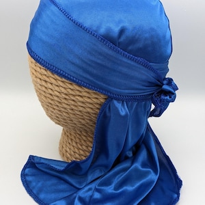 May include: A royal blue satin durag with a long tail. The durag is tied at the back of the head and has a blue stitching around the edges.