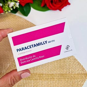 Può includere: Una scatola bianca e rosa di compresse Paracetamilly 360 CFU. La scatola ha un design rettangolare rosa e il testo "Paracetamilly compresse" in blu. La scatola è tenuta da una persona.