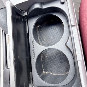 May include: A black plastic car console with two circular cup holders. The word "Solara" is printed on the side of the cup holder. The console is open, revealing the cup holders and a small storage compartment.