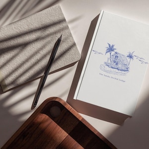 May include: A wooden serving tray, a gray notebook, a black pencil, and a white book with a blue illustration of a beach house and the text "The Sandy Orchid Lodge". The items are arranged on a light-colored surface.