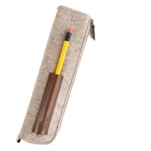 Könnte beinhalten: Ein beigefarbener Filz-Bleistiftetui mit einem braunen Leder-Bleistiftfach und einem Reißverschluss. Das Etui trägt den Schriftzug "The Magic" auf der Seite.