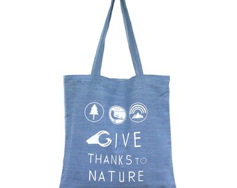 以下が含まれることがあります： 白い文字で「GIVE THANKS TO NATURE」と書かれた青いキャンバス地のトートバッグ。バッグの上部には、木、鳥、山、虹の4つの白いアイコンがあります。
