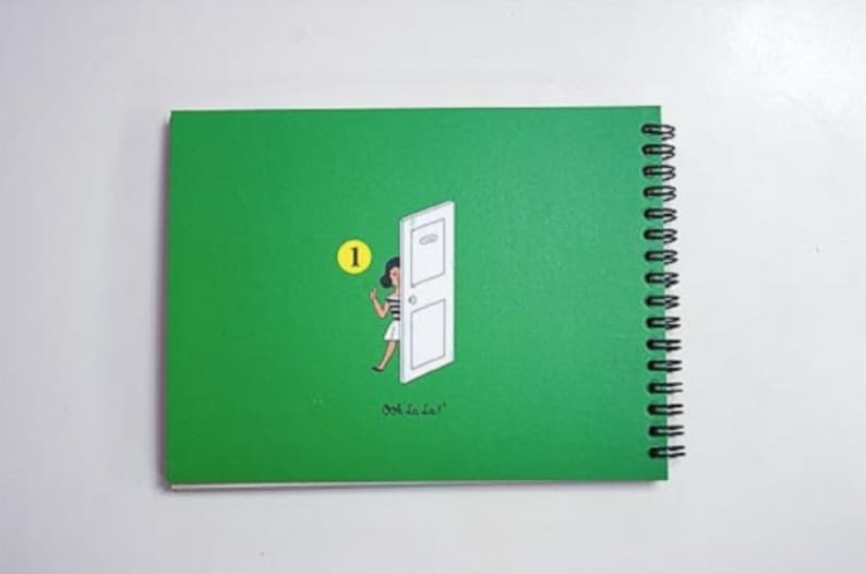 May include: A green spiral bound notebook with a white door and a woman in a white dress and black hair peeking out. The number "1" is circled in yellow. The text "ooh la la" is printed on the cover.