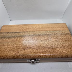 May include: A rectangular wooden box with a natural wood grain finish. The box has a silver-coloured metal clasp and hinges. The wood has a light brown colour with darker brown streaks. The box is closed.