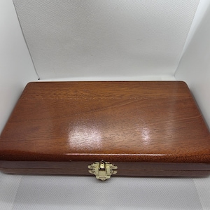May include: A rectangular wooden box with a polished, dark brown finish. The box has a gold-coloured clasp in the centre. The wood grain is visible, and the box appears to be closed.