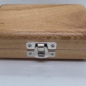 May include: A rectangular wooden box with a light brown finish. The box has a metal clasp in the centre to secure the lid. The wood grain is visible on the surface of the box.