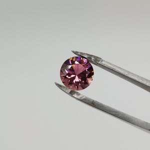Könnte beinhalten: Ein runder, facettierter Edelstein mit einem rosa und violetten Farbton, gehalten von einer Pinzette vor einem weißen Hintergrund.