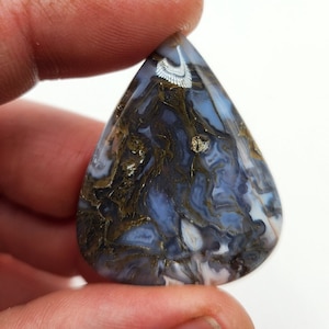 Könnte beinhalten: Ein tropfenförmiger Cabochon aus blauem, weißem und goldenem Achat mit einem wirbelnden Muster. Der Achat hat eine natürliche, polierte Oberfläche.